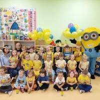 Dzieci ustawione w trzech rzędach pozują do zdjęcia. Z lewej strony Pani, z prawej postać Minionka. Za nimi półki z zabawkami, tablica z obrazkami, na półkach wazony z żółtymi i niebieskimi balonami.