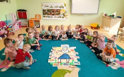 Grupa dzieci siedzi w półkolu na dywanie. W środku półkola plakat z namalowaną Kicią Kocią oraz obrazkami 