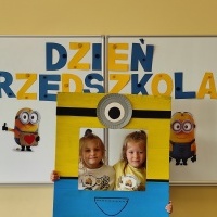 . Dwie dziewczynki stoją w fotobudce z minionkiem. W tle tablica z napisem Dzień Przedszkolaka.