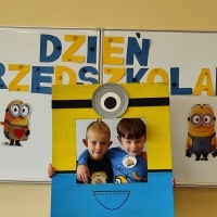 Dwóch chłopców stoi w fotobudce z minionkiem. W tle tablica z napisem Dzień Przedszkolaka.