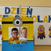 Dwóch chłopców stoi w fotobudce z minionkiem. W tle tablica z napisem Dzień Przedszkolaka.