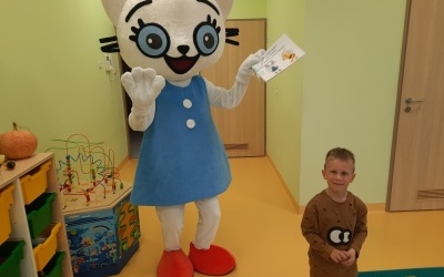 Kicia Kocia trzymająca książeczkę podarowaną przez dzieci. Przed nią z prawej strony chłopiec w brązowym dresie. W tle drzwi. 