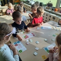 Dzieci siedzące przy stolikach przyklejają naklejki. Z prawej strony w tle jesienny kącik na parapecie.