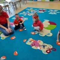 Troje dzieci i Pani ubrani na czerwono, w opaskach z jabłkiem siedzą na dywanie. Dopasowują do siebie połówki jabłek. W tle stoliki z krzesełkami.