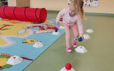 Dziewczynka wykonuje ćwiczenia podnoszenie i osadzanie piłeczki na pachołku sportowym. 