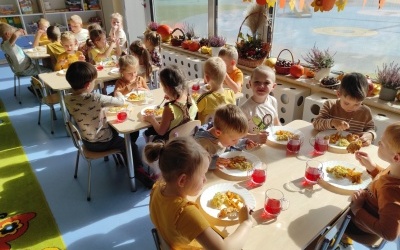 Dzieci siedzą przy stolikach. Jedzą kotleciki i ziemniaki. W tle okno i jesienne ozdoby.