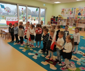 Dzieci ubrane odświętnie, śpiewają hymn polski w tle biało-czerwona flaga.