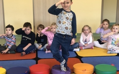 Chłopiec stoi na fioletowym krążku. W języku migowym pokazuje kolor fioletowy. Obok chłopca leżą kolorowe krążki. W tle dzieci siedzą po turecku. 