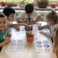Grupa dzieci siedzi przy stoliku, kolorują emotki przedstawiające nastroje. 