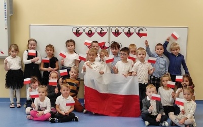 Dzieci ustawione w trzech rzędach trzymają chorągiewki biało-czerwone. W tle napis Kocham Cię Polsko oraz flaga Polski.