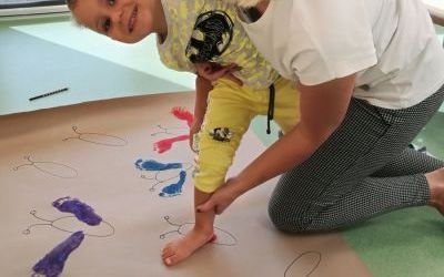 Chłopiec z pomocą nauczycielki odbija pomalowaną stopę na szarym arkuszu papieru.