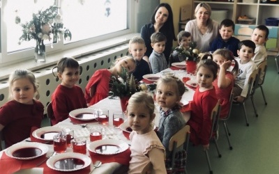 Grupa dzieci wraz z Paniami siedzi przy uroczyście nakrytym stole wigilijnym. W tle okno oraz zimowe i świąteczne dekoracje. 