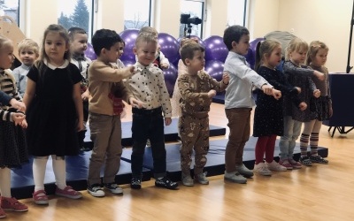 Grupa dzieci stoi w rzędzie na sali gimnastycznej. Dzieci pokazują improwizację ruchową do piosenki śpiewanej Mikołajowi.