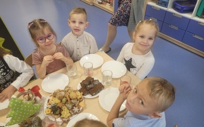 Grupa dzieci siedzi przy stole. Na stole ustawione talerze z ciastkami i dekoracje świąteczne.