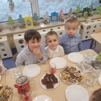 Trzech chłopców siedzi przy wigilijnym stole. Na stole ustawione talerze z ciastkami i dekoracje świąteczne. 