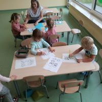 Dzieci siedzą przy dwóch stolikach i kolorują obrazki. W jednym stoliku obok dwóch dziewczynek siedzi nauczycielka. 