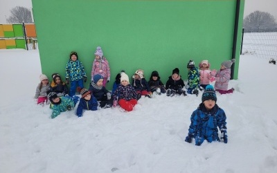 Dzieci siedzą na śniegu. W tle budynek przedszkola.