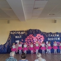 Dziewczynki w białych bluzkach i różowych spódniczkach śpiewają piosenkę dla babci. W tle dekoracja z różowych motylków.