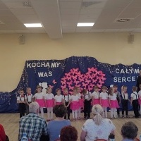 Dzieci ustawione w rzędzie śpiewają piosenki z okazji Dnia Babci i Dziadka. W tle dekoracja z różowych motylków.