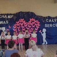 Dzieci ustawione w rzędzie recytują wierszyki z okazji Dnia Babci i Dziadka. W tle dekoracja z różowych motylków.