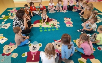 Dzieci wraz z Panią siedzą w kole na dywanie. W środku koła dziewczynka odsłania karty do gry memory.