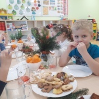 Grupa dzieci siedzi przy uroczyście nakrytym stole wigilijnym. Zjadają ciasteczka świąteczne. W tle półeczki z zabawkami.