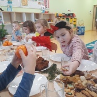 Grupa dzieci siedzi przy uroczyście nakrytym stole wigilijnym. Zjadają ciasteczka świąteczne oraz mandarynki. W tle półeczki z zabawkami.