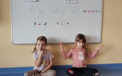 Dziewczynki siedzą na podłodze i pokazują w języku migowym wyrazy: lubię luty.