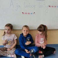 Grupa dzieci siedzi na podłodze i pokazuje w języku migowym wyraz: 