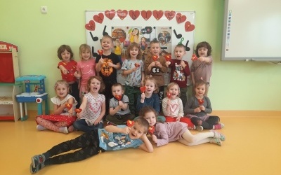 Dzieci ustawione w trzech rzędach. W rękach trzymają lizaki w kształcie serca. Za nimi tablica z napisem Walentynki.