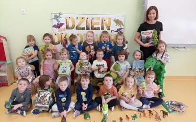 Dzieci wraz z Panią ustawione w trzech rzędach. W rękach trzymają książki i figurki dinozaurów. Za nimi tablica z napisem 
