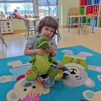 Chłopiec siedzi na dywanie. Trzyma w rękach zielonego, pluszowego dinozaura. Za nim stoliki z krzesełkami oraz okna wychodzące na plac zabaw.