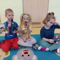 Chłopiec i dwie dziewczynki siedzą na dywanie. W rękach trzymają figurki i pluszaki dinozaurów.
