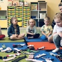 Dzieci wraz z Panią siedzą w kole na dywanie. W środku koła leżą kolorowe skarpetki, dziewczynka szuka pary skarpet.