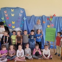 Grupa dzieci wraz z wychowawcą pozuje do zdjęcia z gościem Panią Zuzanną Trojanowską - Dasiak. W tle dekoracja z księgą i motylkami.