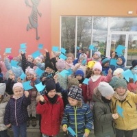 Pszczółki i Kotki zdjęcie wspólne przed budynkiem przedszkola, dzieci trzymają w rękach niebieskie chorągiewki.