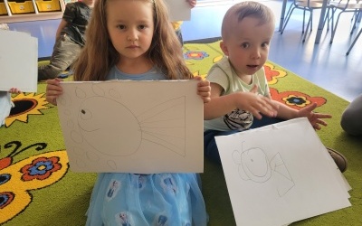 Dziewczynka z chłopczykiem siedzą na dywanie. Pokazują arkusz papieru, na którym samodzielnie narysowali rybę