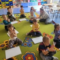 Dzieci siedzą na dywanie przed dużymi arkuszami papieru. Pokazują znak 