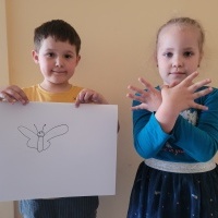 Dwójka dzieci stoją przy ścianie. Chłopiec pokazuje rysunek motyla, a dziewczyna miga znak 