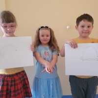 Trójka dzieci stoją przy ścianie. Dwójka dzieci pokazuje rysunek żółwia. Dziewczynka pokazuje znak 