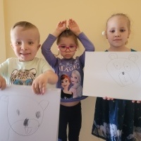 Trójka dzieci stoją przy ścianie. Dwójka dzieci pokazuje rysunek niedźwiadka. Dziewczynka w okularkach pokazuje znak 