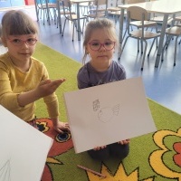Dwie dziewczynki siedzą na dywanie. Jedna dziewczynka pokazuje kartkę, na której narysowała rybę. Druga dziewczynka pokazuję znak 