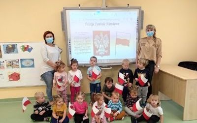 Dzieci trzymają chorągiewki biało -  czerwone. Za nimi  tablica multimedialna z Polskimi Symbolami Narodowymi. Z lewej i z prawej strony tablicy stoją panie.