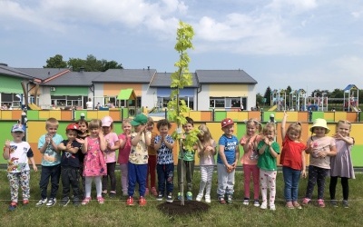 Grupa dzieci pozuje do zdjęcia przy posadzonym drzewie. W tle budynek przedszkolny.