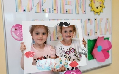 Dwie dziewczynki pozują w ramce z Okazji Dnia dziecka, w tle napis na tablicy magnetycznej.