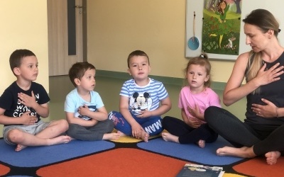 Grupa dzieci siedzi po turecku w kole razem z Panią. Dzieci uczą się praktyki oddechu.