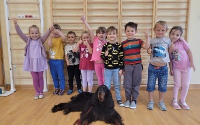 Grupka dzieci stoi przy drabinkach na sali gimnastycznej. Przed nimi leży pies. 