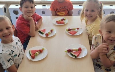 Piątka dzieci siedzi przy stoliku. Na stoliku znajdują się talerze z owocami.