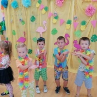 Dzieci w strojach hawajskich tańczą na sali gimnastycznej. W tle kolorowe dekoracje. 