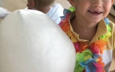 Uśmiechnięty chłopczyk w stroju hawajskim siedzi przy stoliku, w rączce trzyma watę cukrową. 